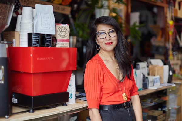 Celeste Wong in a cafe, in front of a La Marzocco espresso machine and coffee merchandise, wearing glasses and a red shirt.